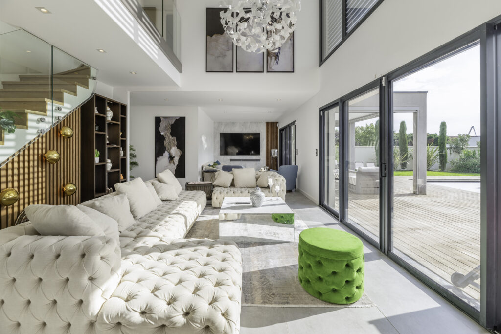 Salon moderne avec de grandes fenêtres, des canapés blancs capitonnés, un pouf en velours vert et des touches de bois. Un escalier avec des appliques murales dorées mène à létage. La pièce souvre sur une terrasse extérieure avec vue sur le jardin. Lustre contemporain et œuvres dart encadrées sur les murs.