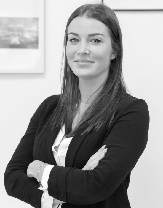 Une femme aux cheveux longs et à l'expression confiante se tient debout, les bras croisés. Elle porte un blazer et un chemisier. L'arrière-plan montre des photos encadrées sur un mur blanc. L'image est en noir et blanc.