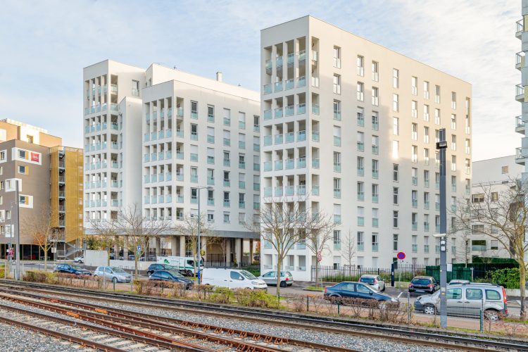 Des immeubles d'appartements blancs modernes aux grandes fenêtres bordent une route remplie de voitures garées, tandis qu'une voie ferrée passe au premier plan. Sur fond de ciel partiellement nuageux, cette scène capture l'essence de Rhône-Alpes avec quelques arbres nus dispersés dans une photographie professionnelle de haute qualité.