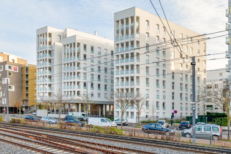 Une scène urbaine moderne dans la région Rhône-Alpes met en scène de hauts immeubles résidentiels blancs avec des balcons. Au premier plan, plusieurs voitures sont garées le long d'une rue avec des voies de tramway parallèles. Des arbres clairsemés et une clôture bordent le trottoir, le tout capturé par une photographie professionnelle de haute qualité.