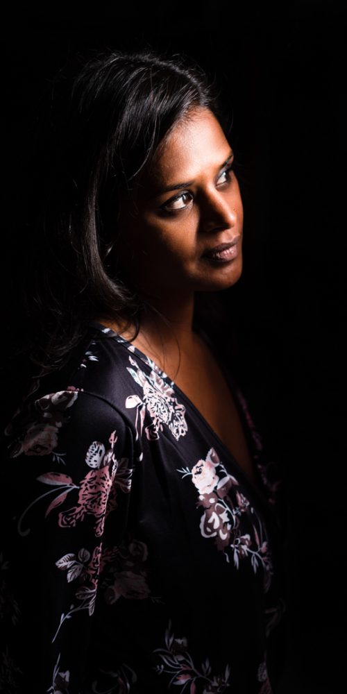 Une femme aux longs cheveux noirs regarde pensivement vers la droite, éclairée par une lumière douce sur un fond sombre. Cette photographie professionnelle de haute qualité de Rhône-Alpes la capture dans une blouse à motifs floraux, renforçant l'atmosphère sereine et contemplative du portrait.