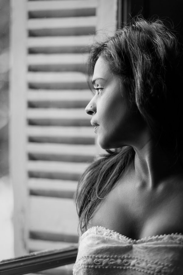 Photographie en noir et blanc d'une femme aux cheveux longs regardant par une fenêtre à volets dans la région Rhône-Alpes. Elle porte un haut bustier et l'image la capture de profil, soulignant son expression contemplative, un véritable exemple de photographie professionnelle de haute qualité.