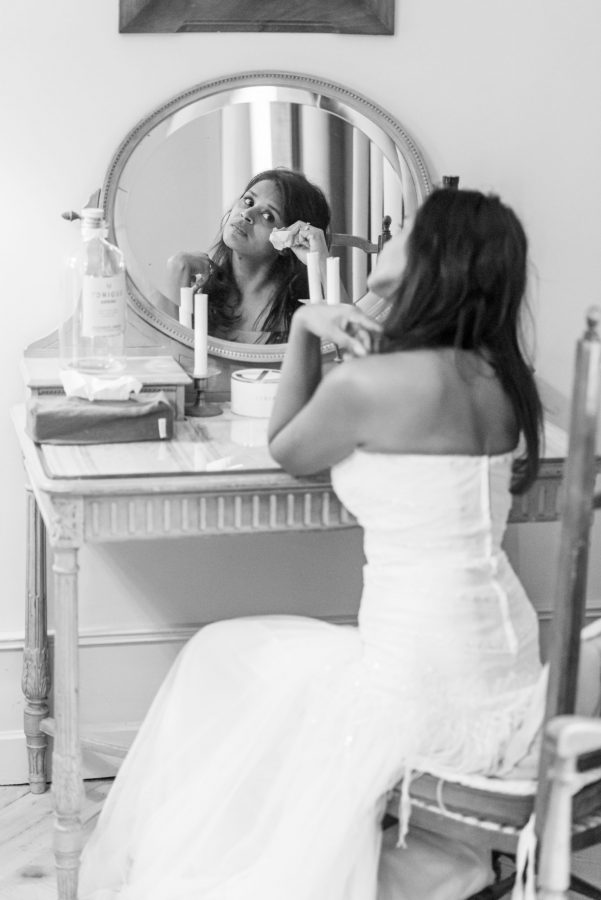 Une femme en robe bustier blanche est assise devant une coiffeuse, les yeux rivés sur le miroir. Elle tient un mouchoir devant son visage dans cette photographie professionnelle de haute qualité. La scène, capturée en noir et blanc, met en valeur des articles de maquillage et une bougie sur la table, évoquant un charme élégant de Rhône-Alpes.