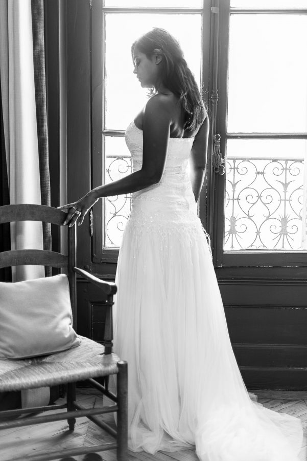 Une photographie professionnelle en noir et blanc de haute qualité montre une femme en robe de mariée bustier debout près d'une fenêtre en Rhône-Alpes. Sa silhouette est magnifiquement mise en valeur alors qu'elle regarde de côté, touchant doucement une chaise d'une main tandis que la lumière entre par derrière.