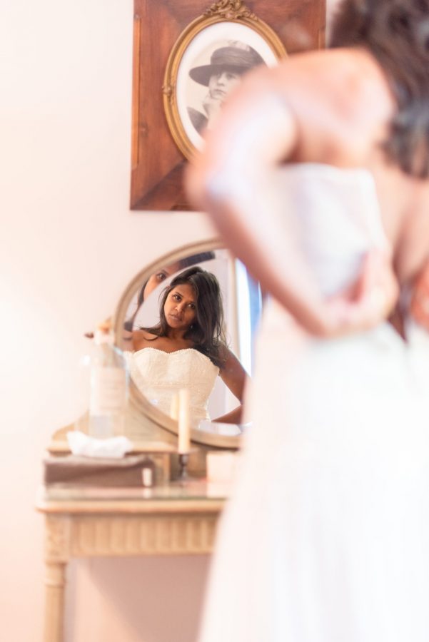 Une femme en robe blanche se reflète dans un miroir rond, ajustant sa tenue dans une pièce à la lumière tamisée. Une photographie aux tons sépia dans un cadre ovale se trouve au-dessus du miroir, représentant une personne portant un chapeau.