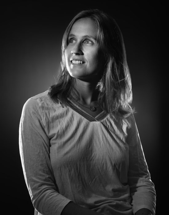 Portrait en noir et blanc d'une femme aux cheveux mi-longs, portant un haut clair. Elle regarde vers le haut avec un léger sourire sur un fond sombre avec un éclairage subtil mettant en valeur ses traits.