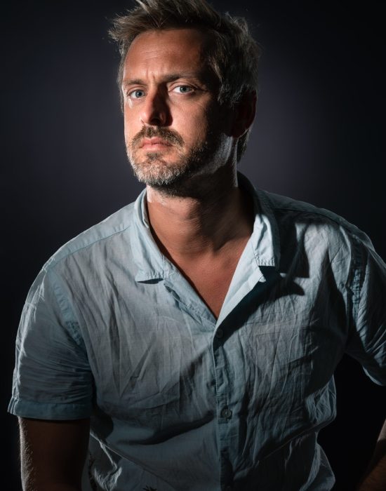 Un homme aux cheveux courts et clairs, portant une barbe et une chemise bleu clair légèrement froissée, est assis dans un décor sombre. Il regarde directement la caméra avec une expression sérieuse, illuminée par une lumière focalisée venant d'en haut.