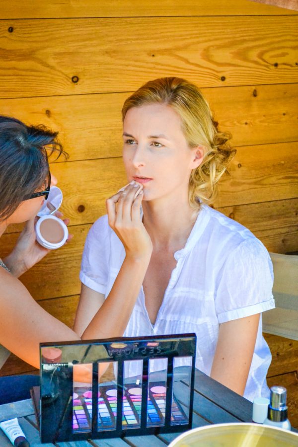 Assise à l'extérieur devant un décor en bois, une femme en chemise blanche se fait maquiller avec brio par une autre femme portant des lunettes de soleil. La scène capture l'essence de la photographie professionnelle de haute qualité, avec une palette de maquillage et des pinceaux visibles au premier plan.