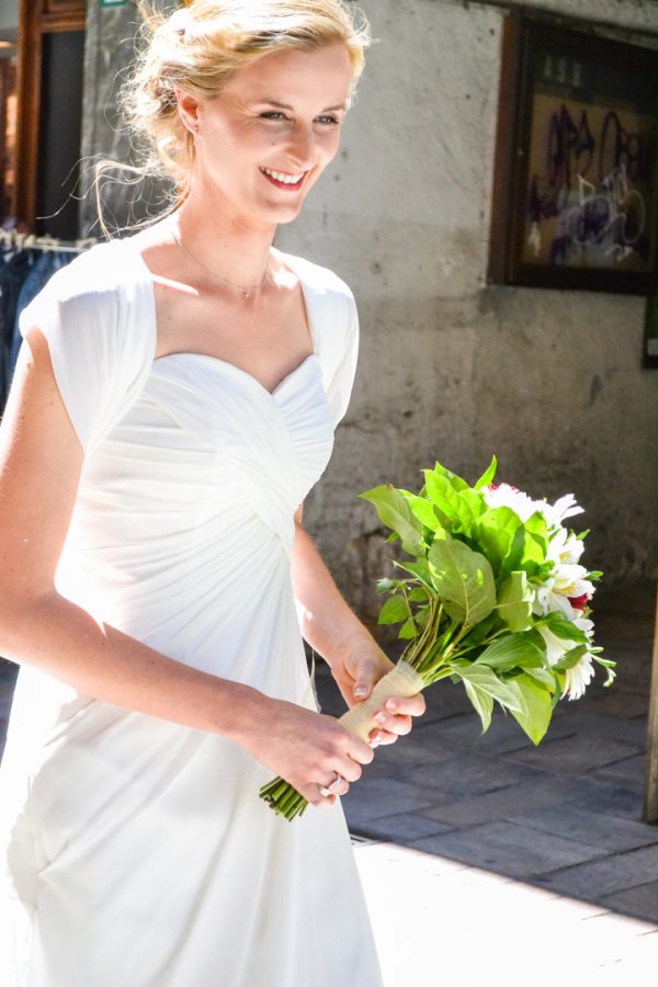 Une femme souriante, vêtue d'une robe de mariée blanche, tient un bouquet de fleurs vertes et blanches. Elle se tient dans une zone urbaine ensoleillée, avec des graffitis sur un mur derrière elle, capturés dans une photographie professionnelle de haute qualité qui met parfaitement en valeur l'esprit vibrant de la région Rhône-Alpes.