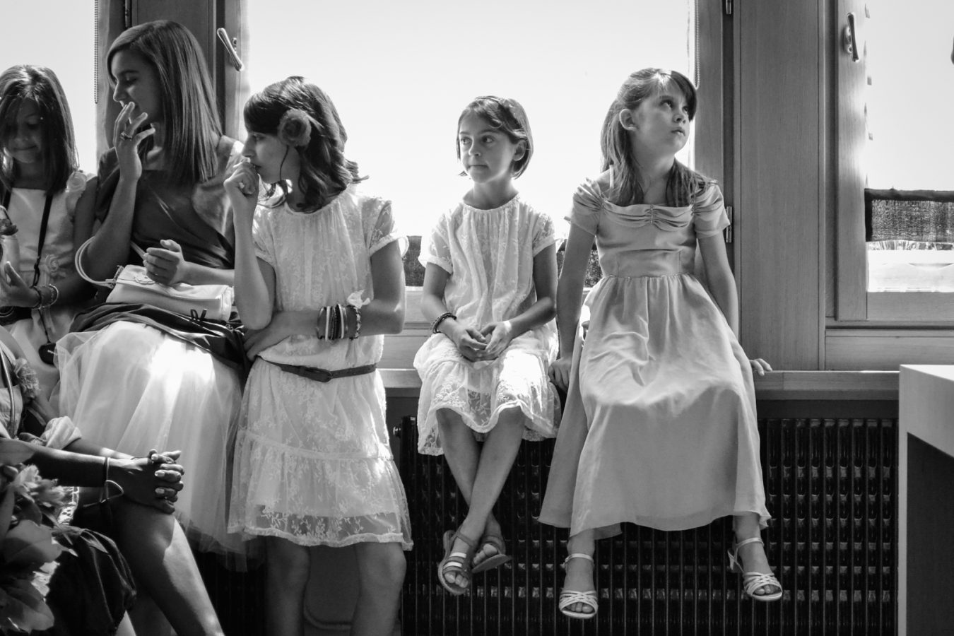 Une photographie en noir et blanc de haute qualité montre cinq jeunes filles assises sur un rebord de fenêtre dans la région Rhône-Alpes. Elles portent des robes et regardent dans des directions différentes, tandis que la lumière du soleil entre élégamment par la fenêtre derrière elles.