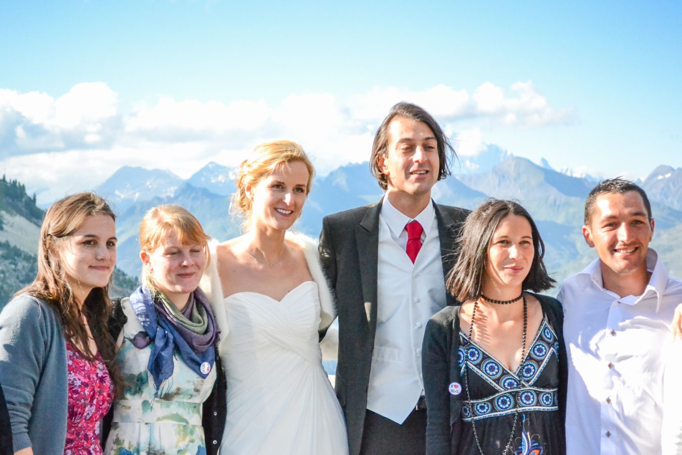 Une mariée en robe blanche et un marié en costume se tiennent avec quatre amis, souriant dans le magnifique décor montagneux de Rhône-Alpes, capturé sous le ciel bleu avec des nuages par une photographie professionnelle haute qualité.