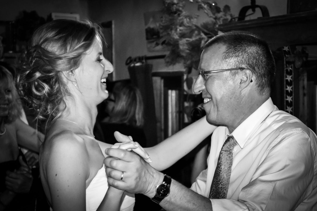 Une photographie professionnelle en noir et blanc de haute qualité capture un couple souriant en train de danser de près lors d'un événement en salle en Rhône-Alpes. La femme, en robe bustier, et l'homme, en chemise et cravate, se tiennent joyeusement la main. D'autres personnes apparaissent doucement floues à l'arrière-plan.