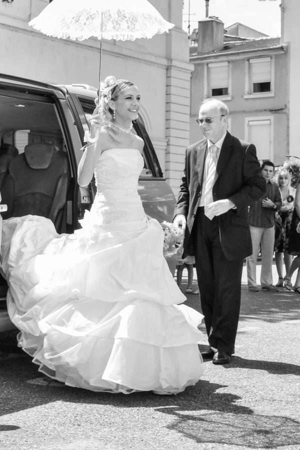 Une mariée en robe blanche sort d'un véhicule, tenant une ombrelle en dentelle, aidée par un homme plus âgé en costume. Une petite foule et des bâtiments sont visibles en arrière-plan. L'image est en noir et blanc.