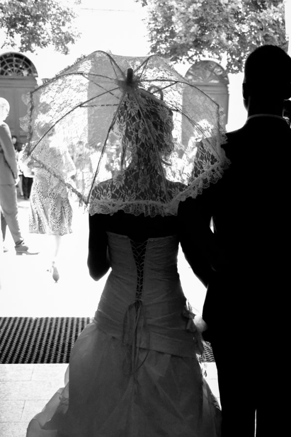 Photographie en noir et blanc d'un couple de mariés réalisée par un photographe professionnel de haute qualité. La mariée tient un parasol en dentelle, debout aux côtés du marié, se détachant sur un paysage extérieur lumineux en Rhône-Alpes. Les invités se mêlent en arrière-plan, tandis que les arbres encadrent élégamment la partie supérieure de l'image.