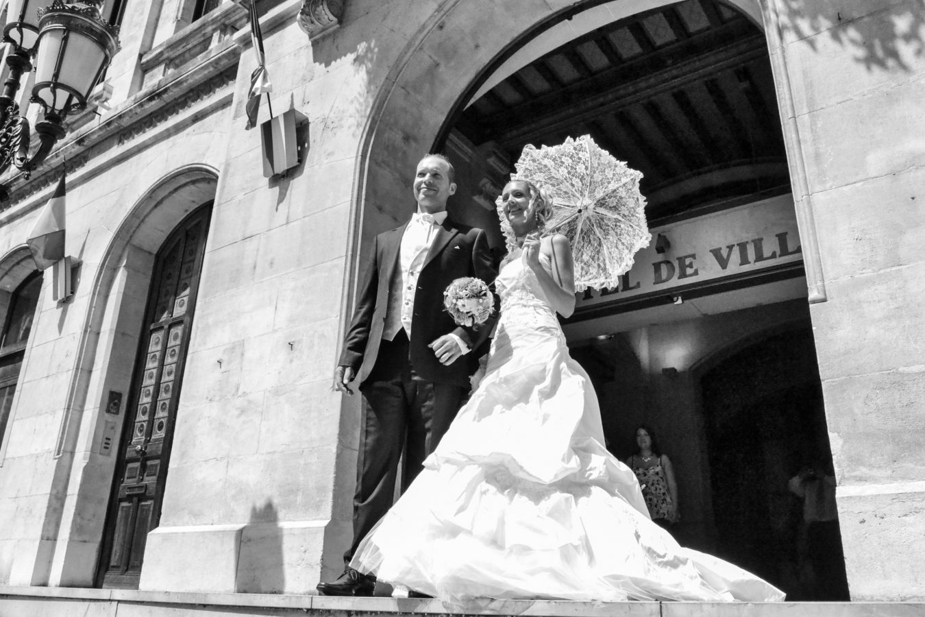 Un couple en tenue de mariage se tient sur les marches d'un bâtiment historique. La mariée tient un parasol en dentelle et un bouquet, tandis que le marié se tient à ses côtés, le tout capturé par une photographie professionnelle de haute qualité. Le cadre ensoleillé de Rhône-Alpes met en valeur l'architecture époustouflante.