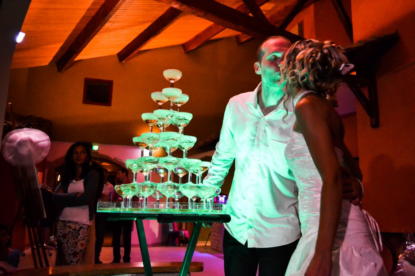 Un couple en tenue de soirée s'embrasse à côté d'une pyramide de flûtes de champagne sous un éclairage chaleureux, capturé avec une photographie professionnelle de haute qualité. Une femme en tenue décontractée observe en arrière-plan. Le décor, avec ses poutres en bois et son haut plafond, évoque le charme de Rhône-Alpes.