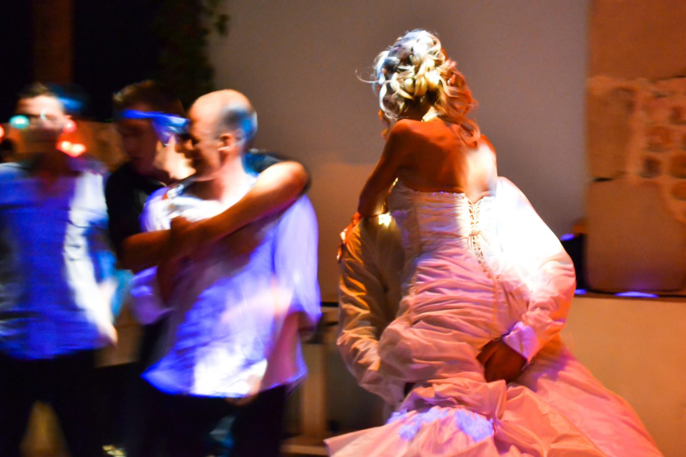 Une scène de réception de mariage dans la région Rhône-Alpes, dans une lumière tamisée, montre une mariée en robe bustier soulevée par un homme. Des silhouettes floues autour d'elles suggèrent des mouvements et des danses, capturées avec une photographie professionnelle de haute qualité, renforçant l'atmosphère festive.