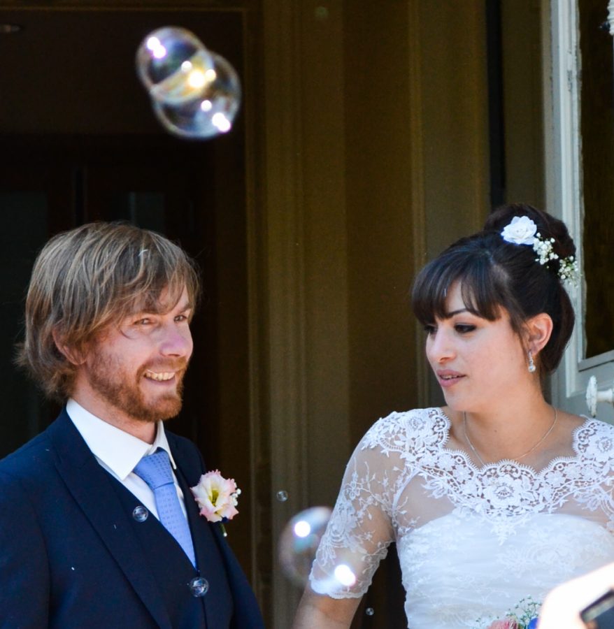 Une mariée et un marié se tiennent ensemble, souriants, dans une photographie professionnelle de haute qualité. La mariée porte une robe de mariée en dentelle et un postiche fleuri, tandis que le marié est en costume avec une boutonnière. Des bulles de savon flottent dans l'air autour d'eux, capturant l'essence enchanteresse de leur célébration Rhône-Alpes.