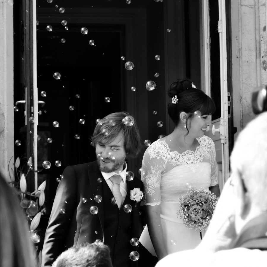 Photographie professionnelle en noir et blanc d'un couple de mariés à l'extérieur d'un bâtiment, entouré de bulles. Dans cette scène Rhône-Alpes de haute qualité, la mariée tient un bouquet dans sa robe en dentelle tandis que le marié se tient élégamment dans son costume.