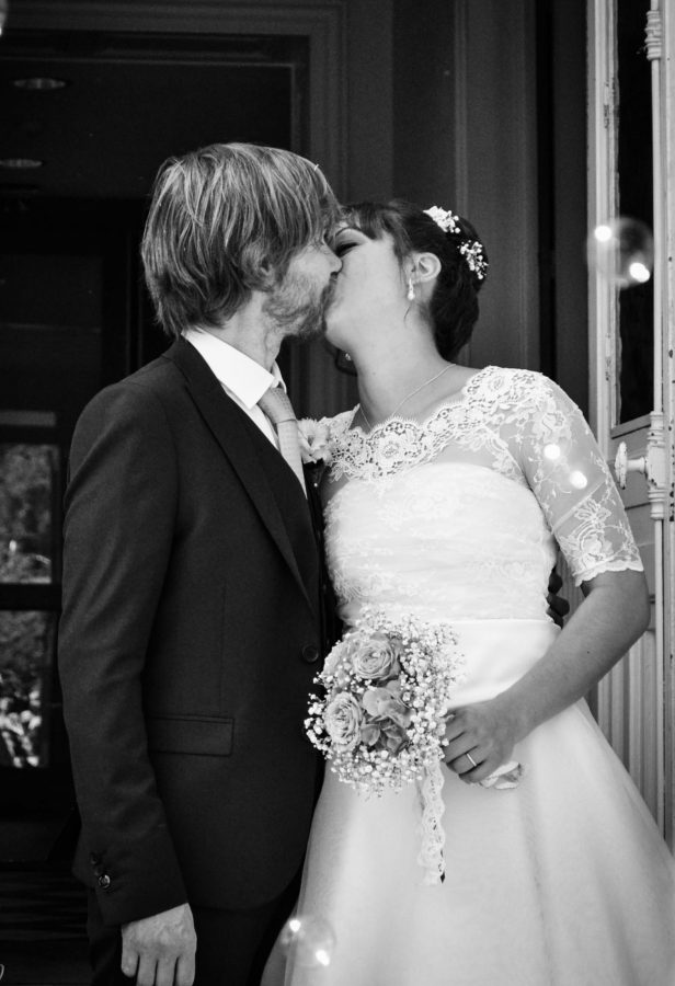 Un couple de mariés s'embrassent le jour de leur mariage. Le marié porte un costume sombre, la mariée éblouit dans une robe blanche aux manches en dentelle, tenant son bouquet. Cette scène en noir et blanc capture l'essence de la photographie professionnelle de haute qualité dans la région Rhône-Alpes.