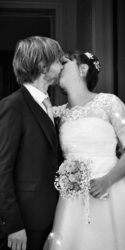 Un couple de mariés s'embrassent le jour de leur mariage. Le marié porte un costume sombre, la mariée éblouit dans une robe blanche aux manches en dentelle, tenant son bouquet. Cette scène en noir et blanc capture l'essence de la photographie professionnelle de haute qualité dans la région Rhône-Alpes.