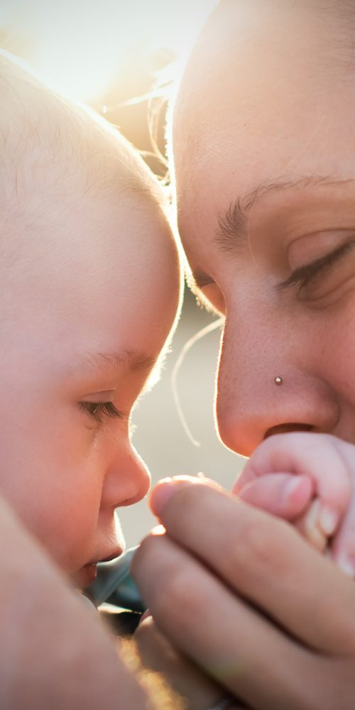 Gros plan d'une femme tenant doucement la main d'un bébé sur son visage, les deux se regardant affectueusement. La lumière du soleil illumine doucement l'arrière-plan, ajoutant une lueur chaleureuse à ce moment tendre.