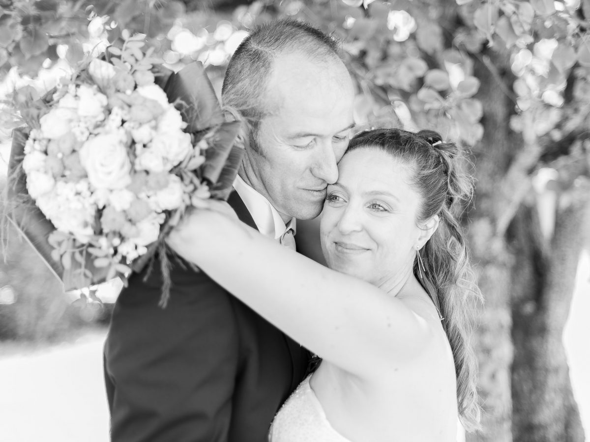 Une photographie professionnelle en noir et blanc de haute qualité montre un couple enlacé sous un arbre. La femme, en robe de mariée, tient un bouquet de fleurs. Tous deux semblent satisfaits et affectueux, l'homme ayant les yeux fermés tandis qu'il se penche vers elle, un moment intemporel de Rhône-Alpes.