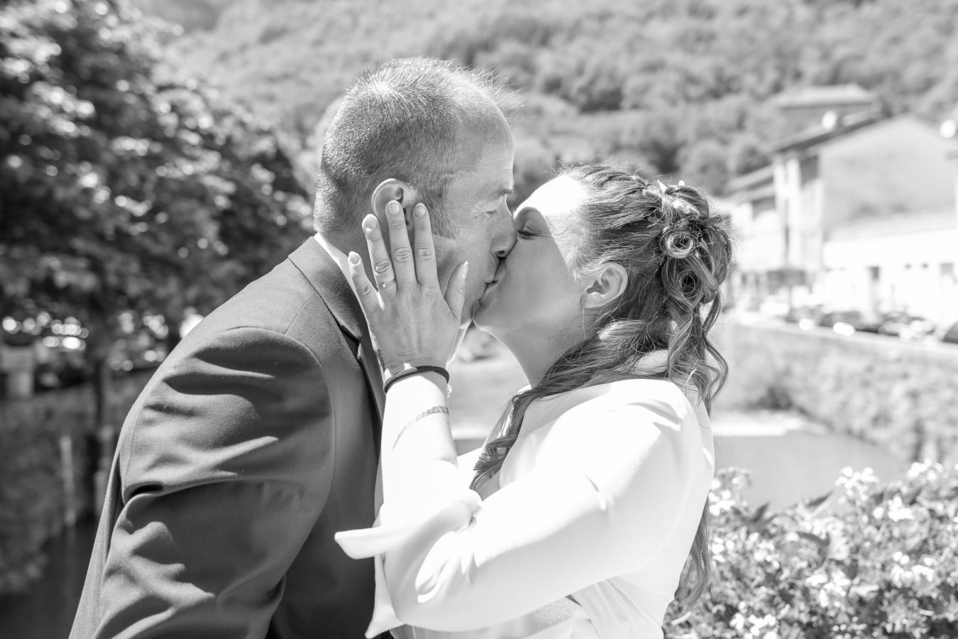 Un couple s'embrasse en plein air par une journée ensoleillée, leur amour capturé avec une photographie professionnelle de haute qualité. L'image en noir et blanc met en valeur le paysage pittoresque de la région Rhône-Alpes, avec ses arbres et ses bâtiments élégants.