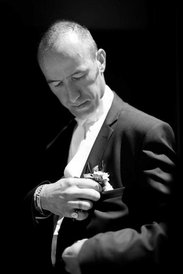 Un homme en tenue de soirée regarde vers le bas tout en ajustant une fleur sur la poche de sa veste de costume. La photographie professionnelle haute qualité en noir et blanc capture son expression contemplative et la texture détaillée de son costume, créant une ambiance élégante et intemporelle dans le décor Rhône-Alpes.