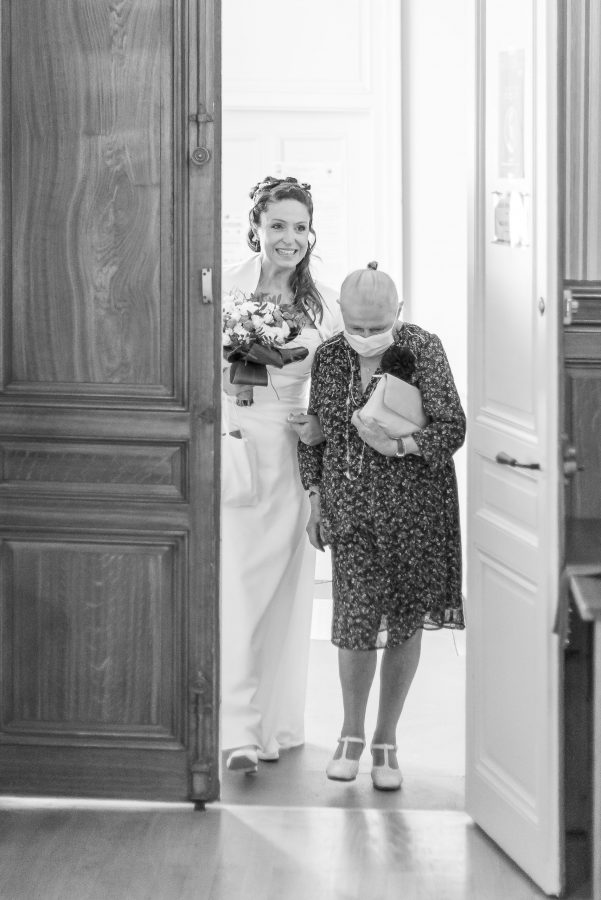 Une mariée en robe blanche et couronne de fleurs entre joyeusement dans une pièce, un bouquet à la main, aux côtés d'une femme âgée en robe à motifs et foulard sur la tête. Capturés par photographie professionnelle haute qualité Rhône-Alpes, leurs sourires illuminent la scène à travers les portes en bois ouvertes.