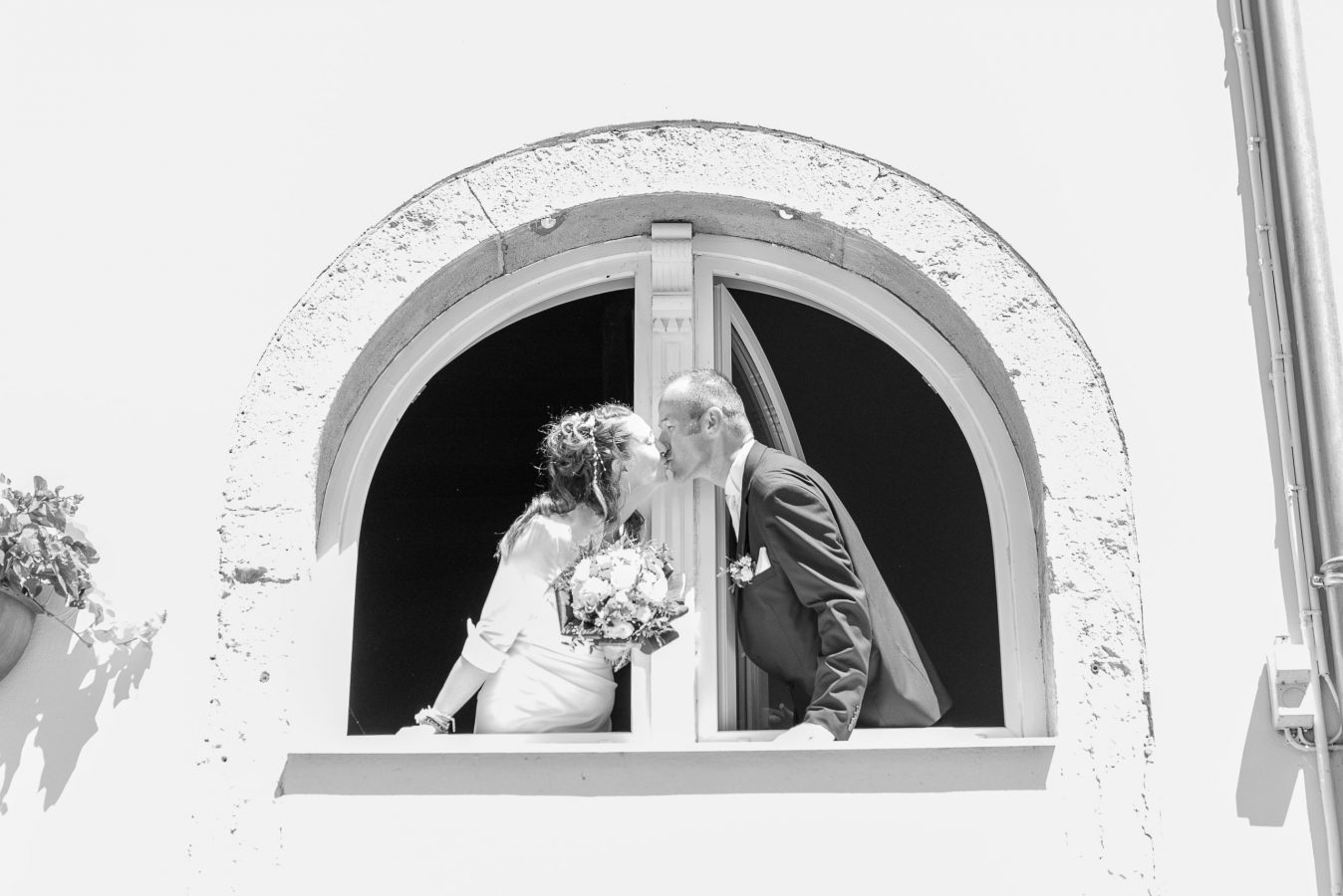 Un couple de mariés s'embrassent en se penchant par une fenêtre en arc, la mariée tenant délicatement un bouquet. Ce moment en noir et blanc capture la romance avec une photographie professionnelle de haute qualité au cœur de Rhône-Alpes.