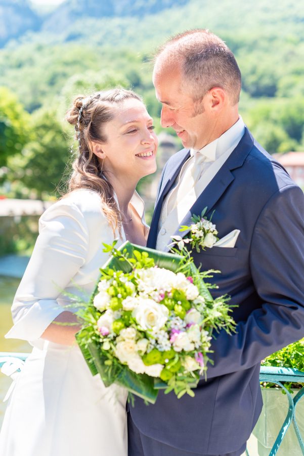 Un couple de mariés se sourient en plein air dans un moment capturé avec une photographie professionnelle de haute qualité. La mariée tient un bouquet de fleurs blanches et vertes alors qu'ils se tiennent sur un balcon surplombant le paysage pittoresque de la région Rhône-Alpes, encadré par une verdure luxuriante et des montagnes majestueuses.