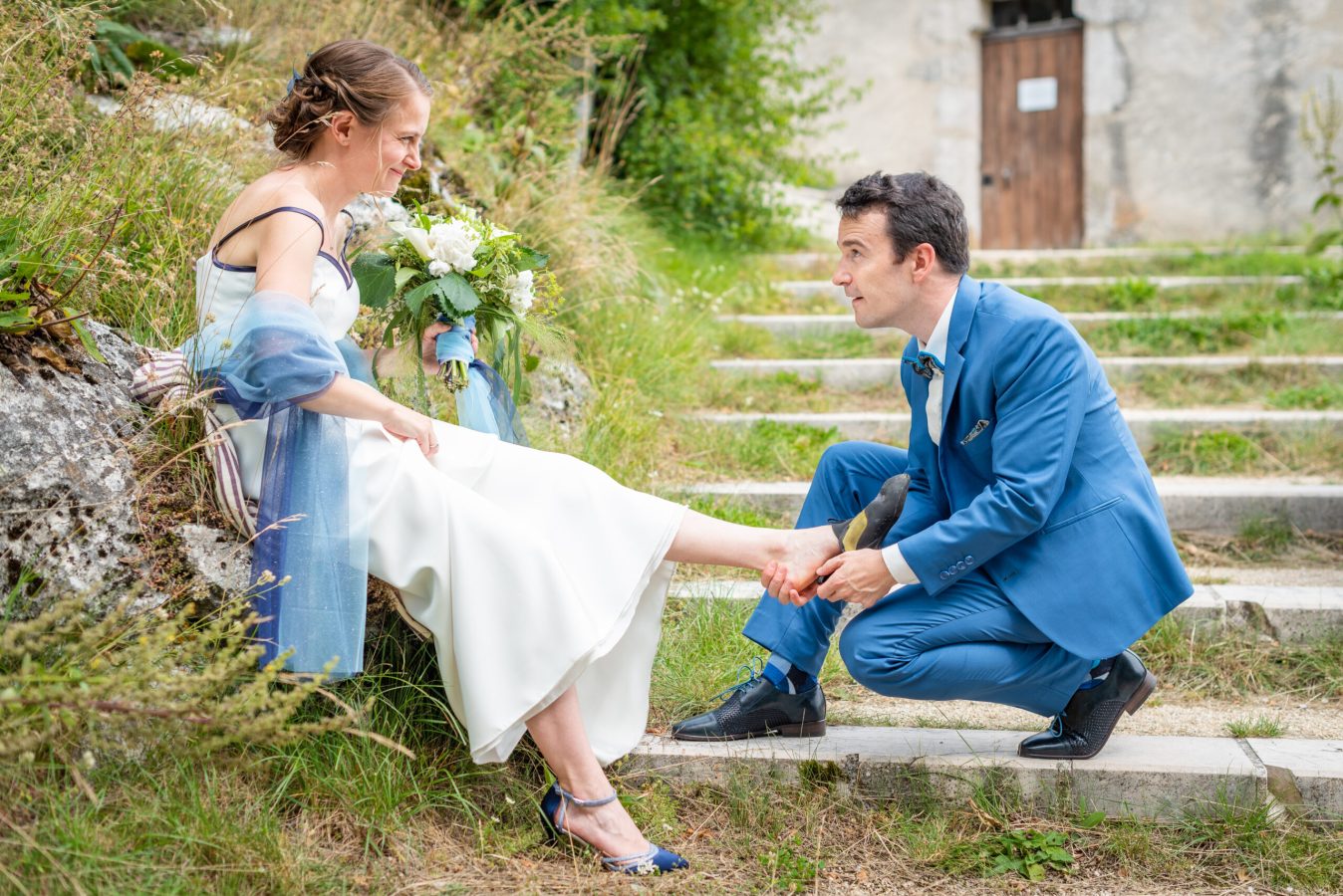 Une mariée est assise sur des marches, tenant un bouquet, tandis quun marié en costume bleu sagenouille et lui met une chaussure au pied. Ils sont à lextérieur, entourés de verdure, avec une porte en bois visible en arrière-plan.