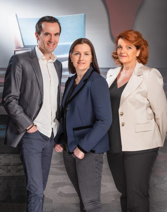 Trois personnes posent pour un portrait, capturé par un photographe expérimenté à Villeurbanne. L'homme à gauche porte un costume gris, la femme au centre arbore un blazer bleu et la femme à droite porte une veste blanche. Derrière eux, des formes abstraites dansent dans une harmonie doucement éclairée.