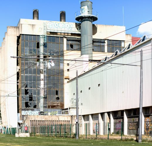 Bâtiment industriel délabré avec vitres cassées, graffitis et structures métalliques capturés dans une photographie professionnelle de haute qualité. Herbe envahissante au premier plan, sous un ciel bleu Rhône-Alpes parsemé de nuages. Des poteaux électriques et des fils électriques complètent la scène.