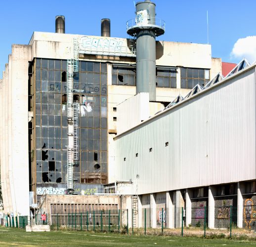 Un grand bâtiment industriel délabré aux fenêtres brisées et aux graffitis sur les murs se dresse majestueusement sous un ciel bleu clair. Avec une haute cheminée et un escalier sur la façade, cette scène capture l'essence de Rhône-Alpes à travers une photographie professionnelle de haute qualité.